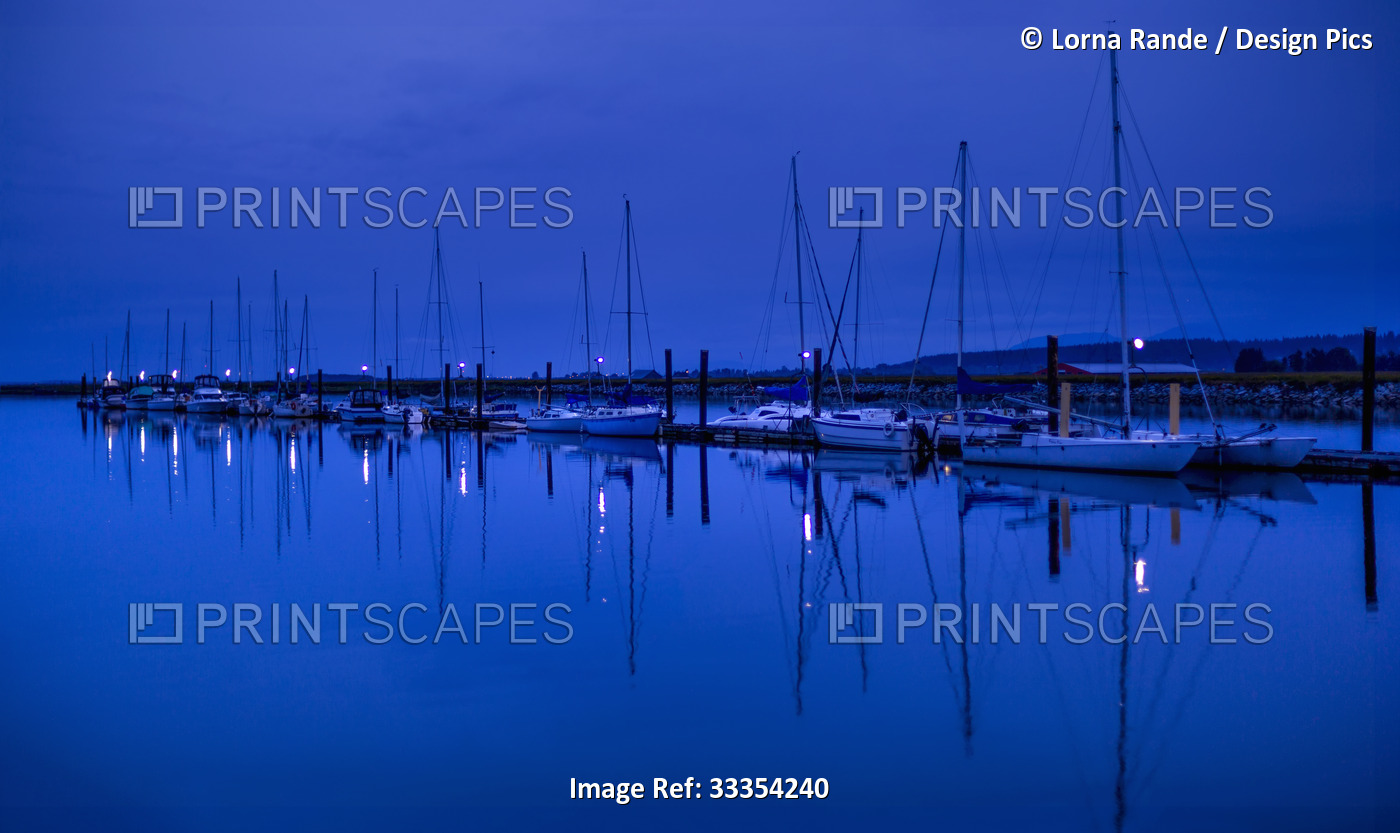 Boats moored in a tranquil harbour at blue hour, with the mirror image of the ...
