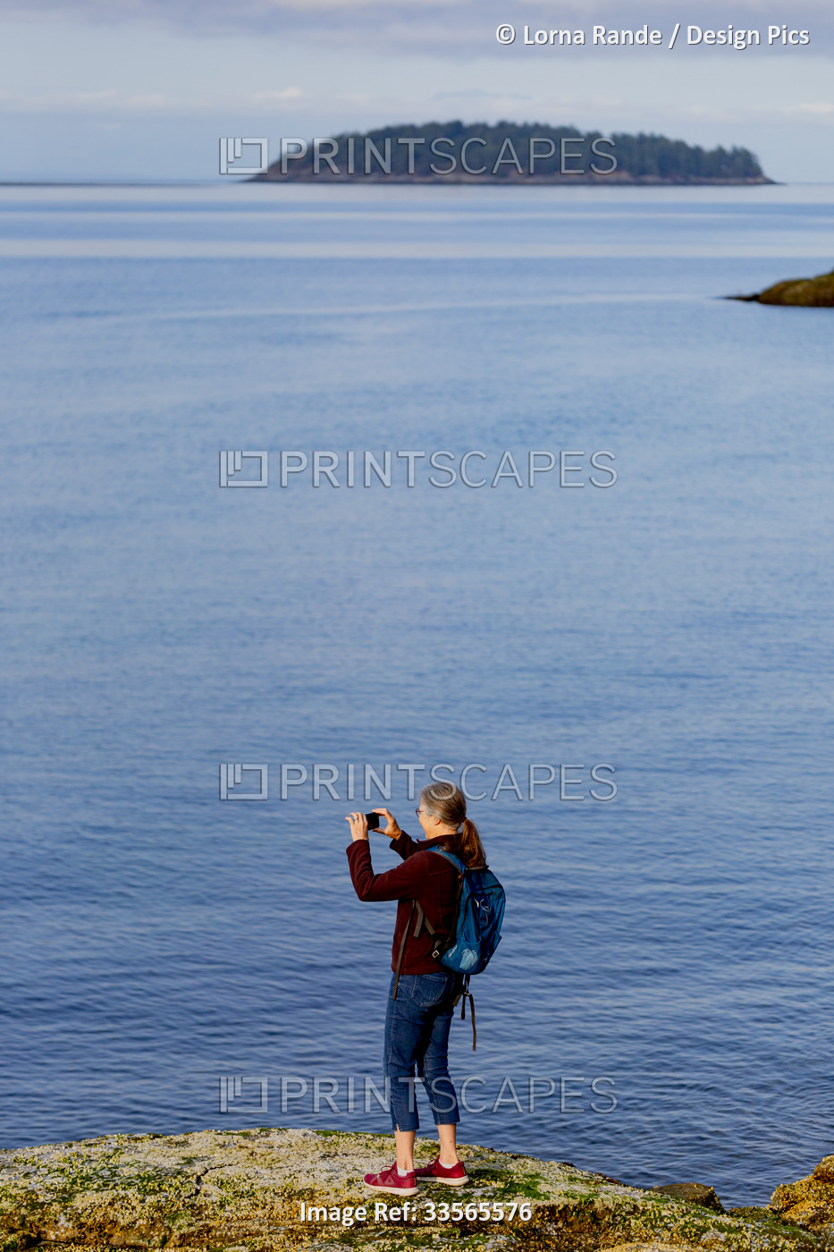 Senior woman stands on a ridge at Sechelt Beach taking a picture of the Pacific ...