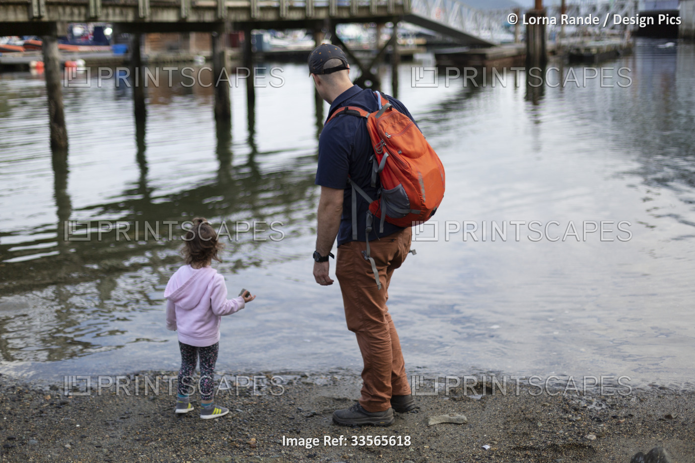 Father and young daughter stand at the water's edge of a beach looking out at ...
