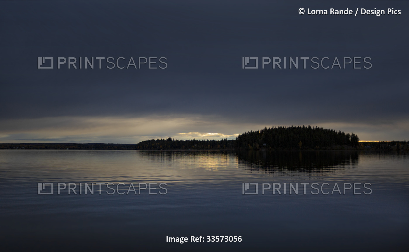 Silhouetted trees along a shoreline and sunset light reflected in the tranquil ...