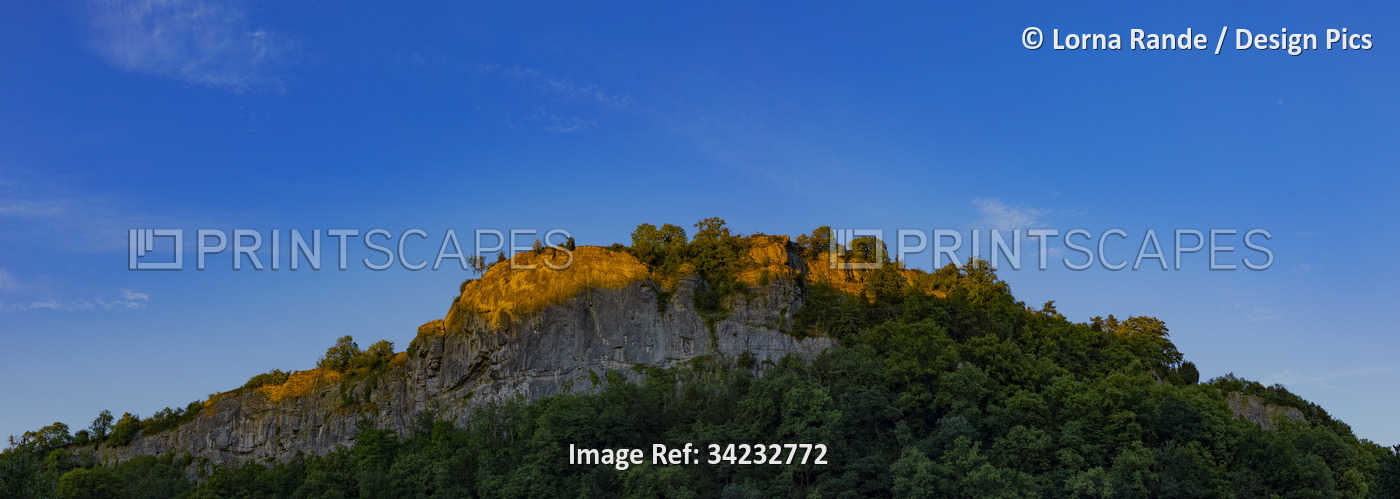Sunlight illuminating top of rocky outcrop against blue sky; Matlock, ...